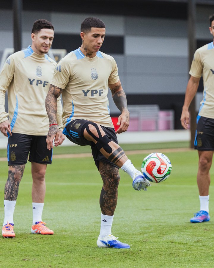 Three male athletes in light blue YPF branded training jerseys and dark blue shorts stand on a grassy field near a covered area with white walls. The central athlete with tattoos on his arms and legs balances on his left leg while kicking a colorful soccer ball with his right foot, wearing a blue knee brace. The other two athletes stand nearby in similar uniforms and white socks with orange cleats.