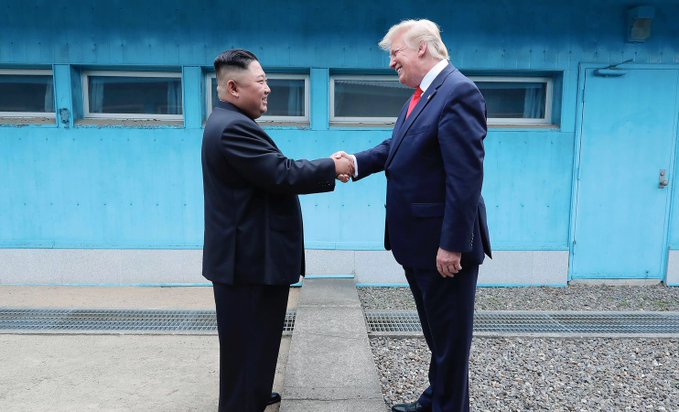 Two men in dark suits shake hands outdoors in front of a light blue building with windows and a door, one with short black hair and the other with light hair wearing a red tie, set against a gravel path and fence.