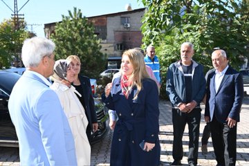 First image shows a large group of about 20 people standing together outside a building with a sign reading Malatya Küçük Sanayi Sitesi, including men and women in business casual attire such as suits, dresses, and headscarves, posing formally on pavement near trees and potted plants. Second image depicts a smaller group of eight individuals outdoors near parked cars and trees, with a woman in a blue coat and scarf gesturing while others in coats and jackets listen attentively. Third image features six people walking on a street lined with trees and buildings, including a woman in a red scarf and blue coat leading, accompanied by men in suits and casual wear. Fourth image portrays an indoor meeting scene with a woman in a blue blazer and red scarf seated at a desk with documents and a salad bowl, flanked by two men in shirts, Turkish and yellow flags on walls, and a large portrait of a man with a quote below.
