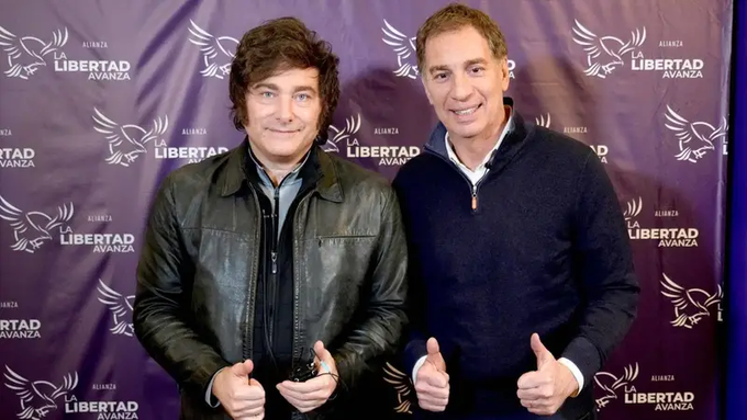 Two men standing side by side in front of a purple backdrop with repeated La Libertad Avanza logos and eagle emblems. The man on the left has curly dark hair, wears a black leather jacket over a blue shirt, holds a small object in his right hand, and gives a thumbs up with his left hand. The man on the right has short light brown hair, wears a dark blue sweater over a collared shirt, and gives thumbs up with both hands.