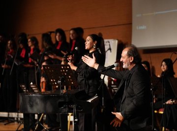First image shows a man in a dark suit speaking at a podium with microphones from TRT Haber and another outlet, against a backdrop with Turkish flag, text Kim Var, and artist name Yücel Arzen Hacıoğulları, in a hall with chairs and Nevşehir Hacı Bektaş Veli University signage. Second image depicts a group of suited men and women including some in headscarves standing around easels with landscape paintings in a modern glass-walled building. Third image captures a woman in black attire singing into a microphone with hands raised, accompanied by a choir of women in red scarves and black outfits, a man in suit holding a microphone nearby, and a piano on stage in a warmly lit auditorium. Fourth image features a large group of people in black attire with red scarves, some holding violins, posing together on stage with a backdrop of Turkish flag and text about warm family artist Yücel Arzen Hacıoğulları, including military uniform and flowers.