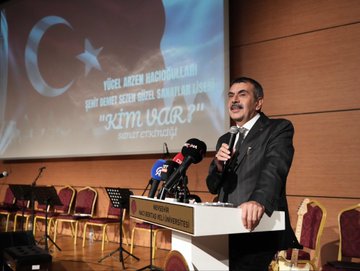 First image shows a man in a dark suit speaking at a podium with microphones from TRT Haber and another outlet, against a backdrop with Turkish flag, text Kim Var, and artist name Yücel Arzen Hacıoğulları, in a hall with chairs and Nevşehir Hacı Bektaş Veli University signage. Second image depicts a group of suited men and women including some in headscarves standing around easels with landscape paintings in a modern glass-walled building. Third image captures a woman in black attire singing into a microphone with hands raised, accompanied by a choir of women in red scarves and black outfits, a man in suit holding a microphone nearby, and a piano on stage in a warmly lit auditorium. Fourth image features a large group of people in black attire with red scarves, some holding violins, posing together on stage with a backdrop of Turkish flag and text about warm family artist Yücel Arzen Hacıoğulları, including military uniform and flowers.