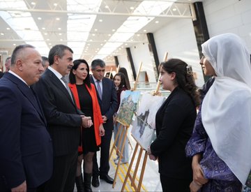 First image shows a man in a dark suit speaking at a podium with microphones from TRT Haber and another outlet, against a backdrop with Turkish flag, text Kim Var, and artist name Yücel Arzen Hacıoğulları, in a hall with chairs and Nevşehir Hacı Bektaş Veli University signage. Second image depicts a group of suited men and women including some in headscarves standing around easels with landscape paintings in a modern glass-walled building. Third image captures a woman in black attire singing into a microphone with hands raised, accompanied by a choir of women in red scarves and black outfits, a man in suit holding a microphone nearby, and a piano on stage in a warmly lit auditorium. Fourth image features a large group of people in black attire with red scarves, some holding violins, posing together on stage with a backdrop of Turkish flag and text about warm family artist Yücel Arzen Hacıoğulları, including military uniform and flowers.
