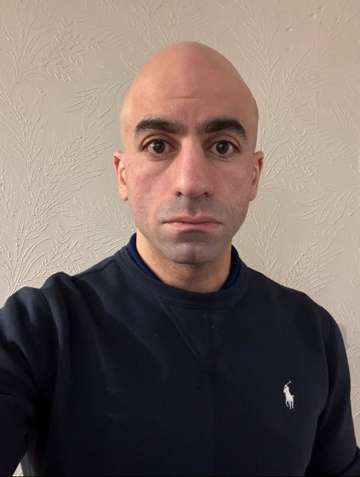 A bald man with dark eyes and a serious expression wears a dark blue hoodie with a small white horse logo on the chest, standing against a plain beige wall in a well-lit indoor setting.