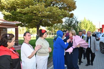 First image shows a group of people including a blonde woman in a red scarf receiving a large bouquet of pink and white flowers from a woman in purple headscarf, with others smiling and standing nearby on a paved area near trees and buildings. Second image depicts a blonde woman in blue dress interacting with women in white uniforms and headscarves under autumn trees by a red building. Third image features a blonde woman holding a pink bouquet while talking with elderly women in headscarves and a man in suit outdoors near a fence and trees. Fourth image captures a group hug involving a blonde woman and several women in headscarves and colorful attire near a building entrance with a lamppost and parked cars.