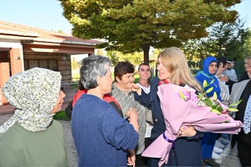 First image shows a group of people including a blonde woman in a red scarf receiving a large bouquet of pink and white flowers from a woman in purple headscarf, with others smiling and standing nearby on a paved area near trees and buildings. Second image depicts a blonde woman in blue dress interacting with women in white uniforms and headscarves under autumn trees by a red building. Third image features a blonde woman holding a pink bouquet while talking with elderly women in headscarves and a man in suit outdoors near a fence and trees. Fourth image captures a group hug involving a blonde woman and several women in headscarves and colorful attire near a building entrance with a lamppost and parked cars.