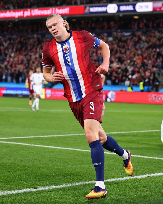Erling Haaland runs on a green soccer field during a match, wearing a red Norway national team jersey with white and blue stripes, number 9 on the back, red shorts with white and blue stripes, blue socks, and yellow Nike cleats. Stadium stands filled with fans in the background, Norwegian flags visible, scoreboard showing UEFA Nations League, sponsor logos on banners.
