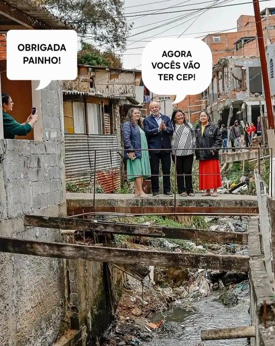 Group of four people stands on a wooden bridge over a polluted stream filled with trash in a favela neighborhood with corrugated metal houses, exposed wiring, and urban decay. A man wearing glasses and a suit jacket poses in the center flanked by three women in dresses and jackets. One man in green takes their photo from the side. Overlay text in white bubbles reads Brigada Agora Obrigada above the group and Votou Cep below near the stream.