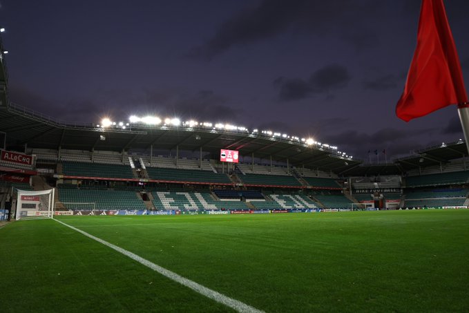 Modern football stadium at night under cloudy skies with illuminated green pitch marked by white lines, empty green spectator stands, large curved roof structure, digital scoreboard showing numbers like 2 and 0, advertising banners on walls, red flag waving on left side, goalposts visible on field ends