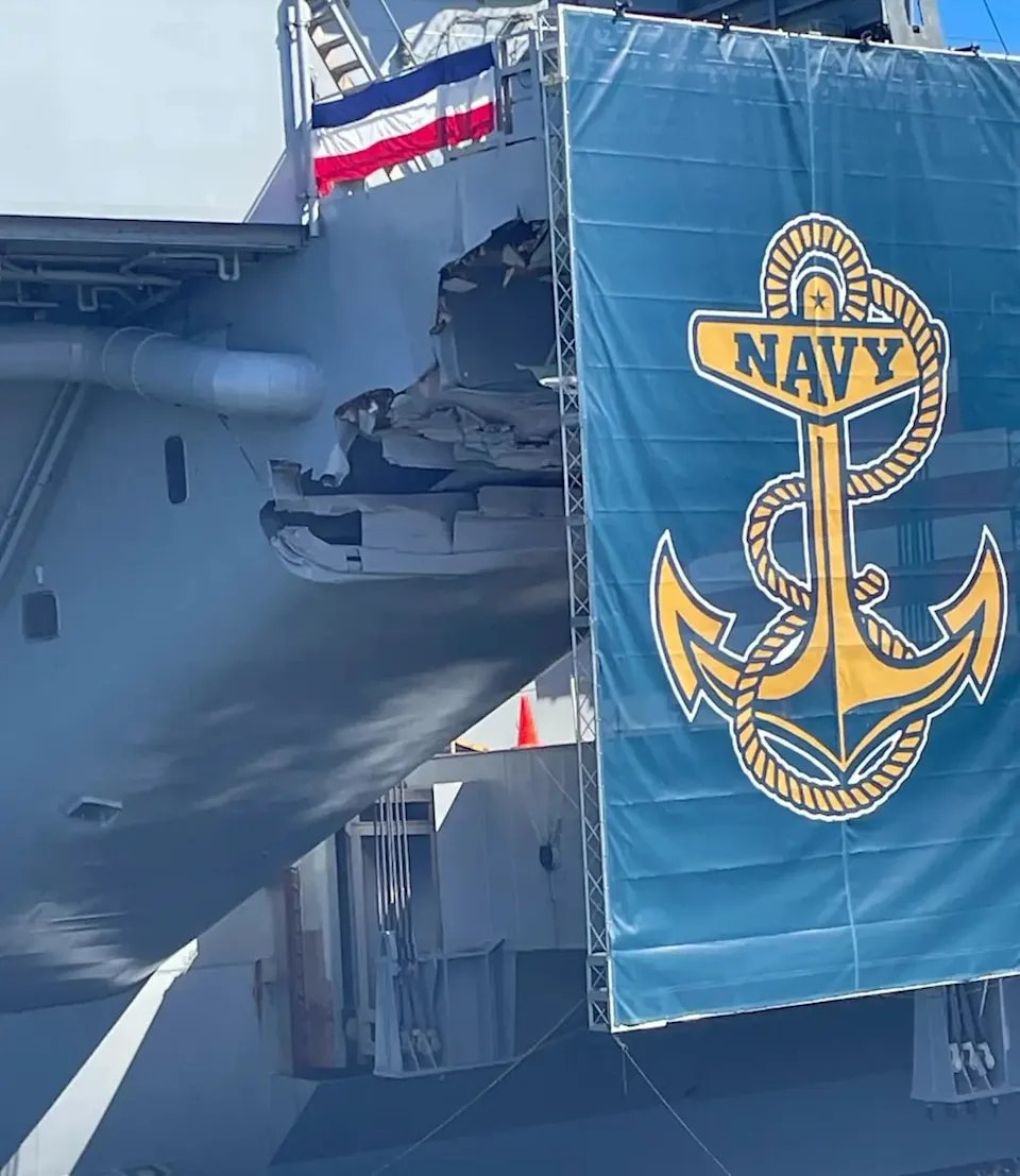Aircraft carrier with gray hull shows structural damage including a large hole and dents on the side near the deck. A large blue navy flag with yellow anchor emblem covers part of the damaged area. Red and white striped banners hang nearby. The ship structure includes railings pipes and metal framework. Background features clear sky and additional ship elements.