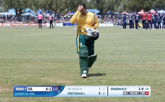 Quinton de Kock in yellow shirt and green pants walks on green cricket field holding helmet, with scoreboard showing Namibia vs South Africa, de Kock 1 off 4 balls, Erasmus 1 off 4, Pretorius 2 overs for 2 runs, crowd and tents in background.