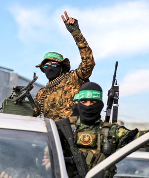 First image shows a man in a blue pinstripe suit, white shirt, and blue tie speaking into two microphones at a podium, with two red Turkish flags on green backdrop behind him, and a small American flag pin on his lapel. Second image depicts two masked men in camouflage clothing and green headbands with Arabic text, one raising a hand in a V-sign while holding a rifle, the other gripping a rifle, standing on a white vehicle with a mounted gun in a sunny outdoor setting with scattered vehicles.