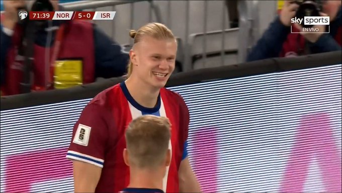 Erling Haaland, a tall blonde player with a ponytail, stands smiling on a football field wearing a red jersey with white accents and the number 9 on his back, surrounded by stadium barriers and photographers, with a scoreboard showing 71 minutes and 5-0 score in the background, and a Sky Sports watermark visible.