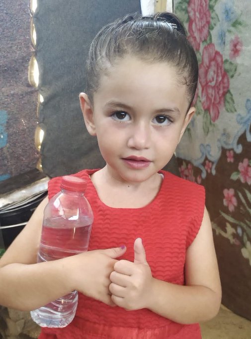 A young girl, Maryam, wearing a red sleeveless dress, holding a plastic bottle of water in one hand and giving a thumbs-up with the other. She has dark hair tied back in a ponytail. The background includes a floral-patterned fabric and a dark surface.