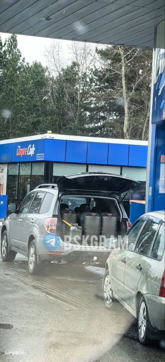 A wet road with a line of cars, including a silver SUV and a sedan, waiting at a gas station. The gas station has a blue building with 