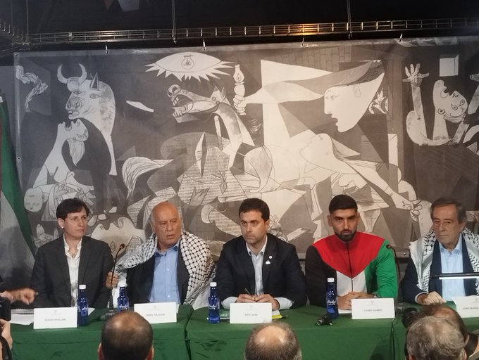 Five individuals seated at a green table with water bottles and nameplates. Behind them, a large black-and-white mural with abstract human and animal figures, including a bull and raised arms, is visible on a backdrop. A green, white, and red flag with a design resembling the Palestinian flag is on the left side. The setting appears to be an indoor event at Gernikako Bakearen museoan.