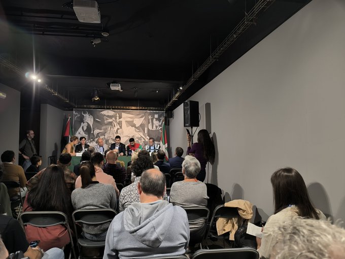 A room with an audience seated on chairs facing a panel of people at a table. A large black-and-white mural with figures and flags, including the Palestinian flag, is visible on the wall behind the panel. The setting appears to be an indoor event space with spotlights and a speaker on the side.