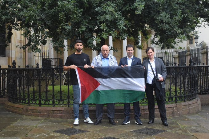 Four men standing outdoors holding a Palestinian flag in front of a building with columns and a black fence. Five men standing indoors in front of a backdrop with "Bakea | Paz" text and a mural. Seven people seated at a table indoors with water bottles, papers, and a backdrop featuring the Palestinian flag and a mural.