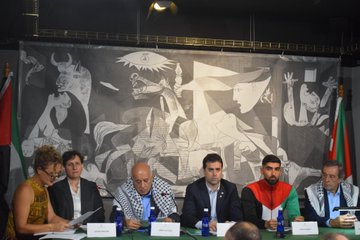 Four men standing outdoors holding a Palestinian flag in front of a building with columns and a black fence. Five men standing indoors in front of a backdrop with "Bakea | Paz" text and a mural. Seven people seated at a table indoors with water bottles, papers, and a backdrop featuring the Palestinian flag and a mural.