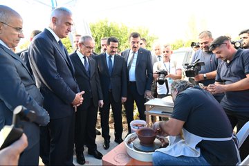 Mehmet Nuri Ersoy and other individuals in suits stand observing a person shaping clay on a pottery wheel. Mehmet Nuri Ersoy and others in suits examine artifacts displayed in glass cases. Mehmet Nuri Ersoy and others in suits view a painting on a wall. A group of people, including Mehmet Nuri Ersoy, sit in chairs at an outdoor event with cameras and banners in the background.