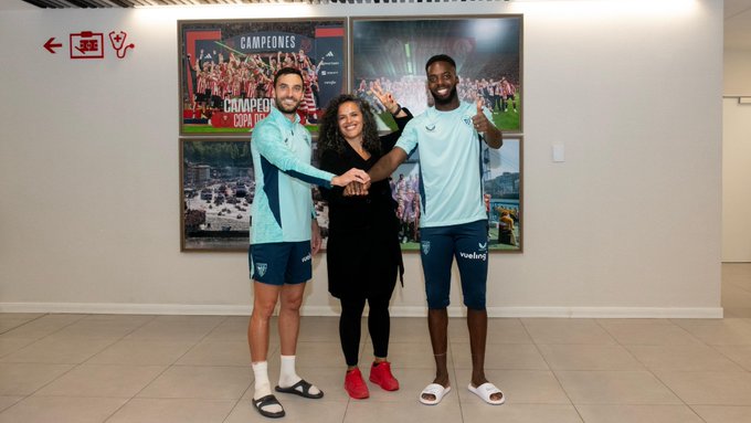 Honey Thaljieh stands in the center, wearing a black outfit and red shoes, linking arms with two men in light blue Athletic Club uniforms and white slippers. They are indoors, in front of a wall with posters displaying Athletic Club imagery, including a "Campeones" title and team photos.
