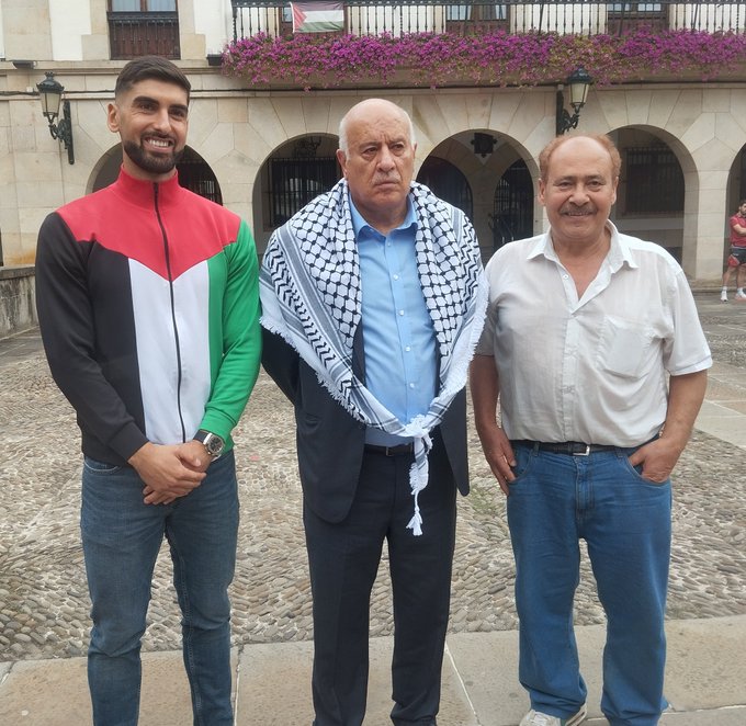 Yaser Hamed wearing a jacket with black, white, red, and green colors. Jibril Rajoub wearing a blue shirt and a black and white checkered scarf. Yaser Hamed\'s father wearing a white shirt and blue jeans. They stand on a cobblestone area in front of a building with arches, balconies, and purple flowers.