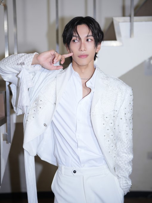 Ikezaki Rihito stands in a white outfit, wearing a white buttoned shirt, white pants, and a white jacket adorned with metallic studs. His dark hair is styled neatly, and he poses with one hand touching his face. The background features a modern indoor setting with stairs and lighting fixtures.