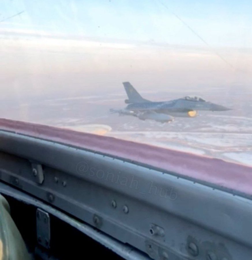从驾驶舱内看到一架 F-16 战斗机并肩飞行。驾驶舱框架和窗户清晰可见，喷气式飞机位于外面，背景是朦胧的天空。存在显示为“@sontah_jr”的水印。