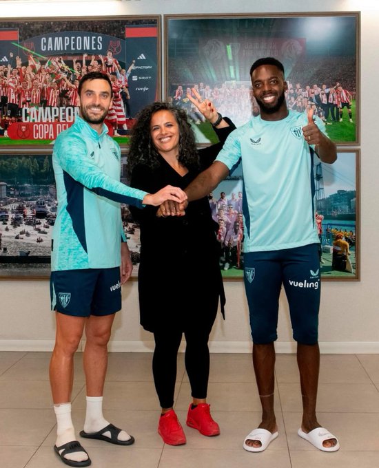 Iñigo Lekue and Iñaki Williams wearing light blue Athletic Club jerseys and shorts, standing in sandals. Honey Thaljieh in a black outfit and red shoes stands between them, shaking hands with both. Background features framed photos of a football team celebrating with a "Campeones Copa del Rey" banner.