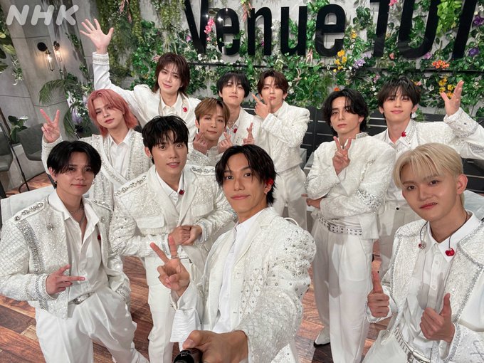 INI members wearing white, embellished outfits with red accents, posing together in a studio setting. They make peace signs and wave, standing in front of a backdrop with greenery and the text "Venue101" visible. An NHK watermark is present in the top left corner.