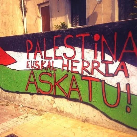 A mural on a wall featuring the word "PALESTINA" in large red letters, with "EUSKAL HERRIA ASKA TU!" below it. The design includes green, white, and red colors resembling the Palestinian flag. The wall is part of an outdoor setting with a tiled sidewalk and a building in the background.