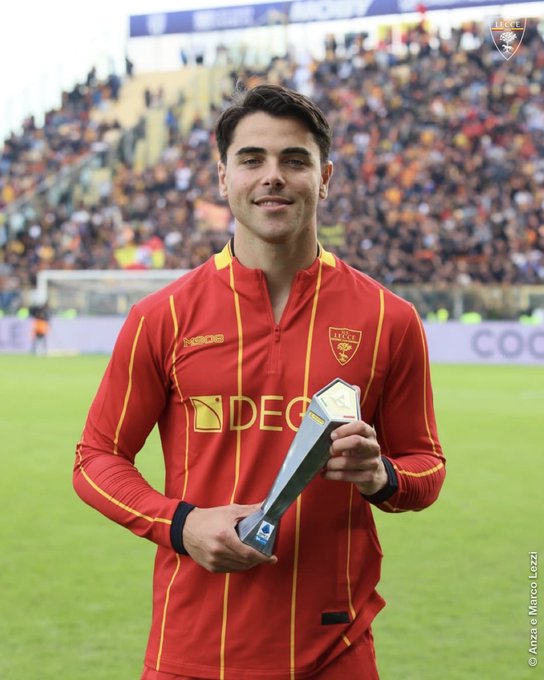 Riccardo Sottil wearing a red and yellow striped soccer jersey with the U.S. Lecce logo, holding a silver trophy on a grassy field. A crowd is visible in the background, seated in stadium stands.