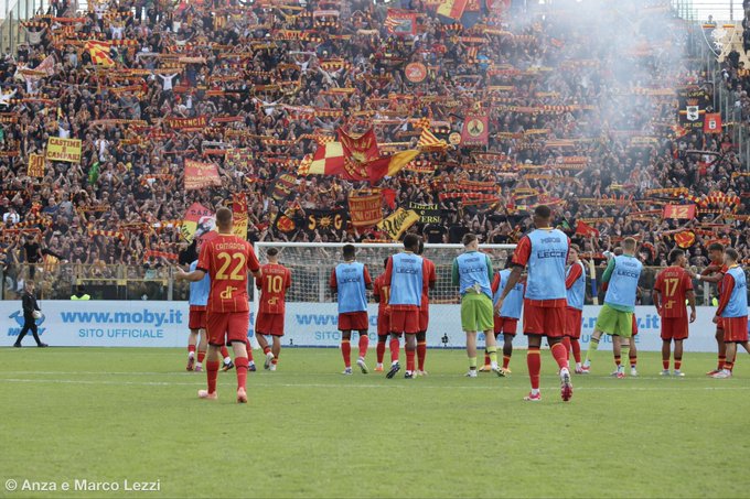 Soccer players in red and blue uniforms stand on a grassy field. Some wear red jerseys with numbers like 22 and 17, while others wear light blue training tops with "www.moby.it" text. A large crowd in the stands holds red and yellow flags, with smoke and banners visible. A goalpost is in the background.