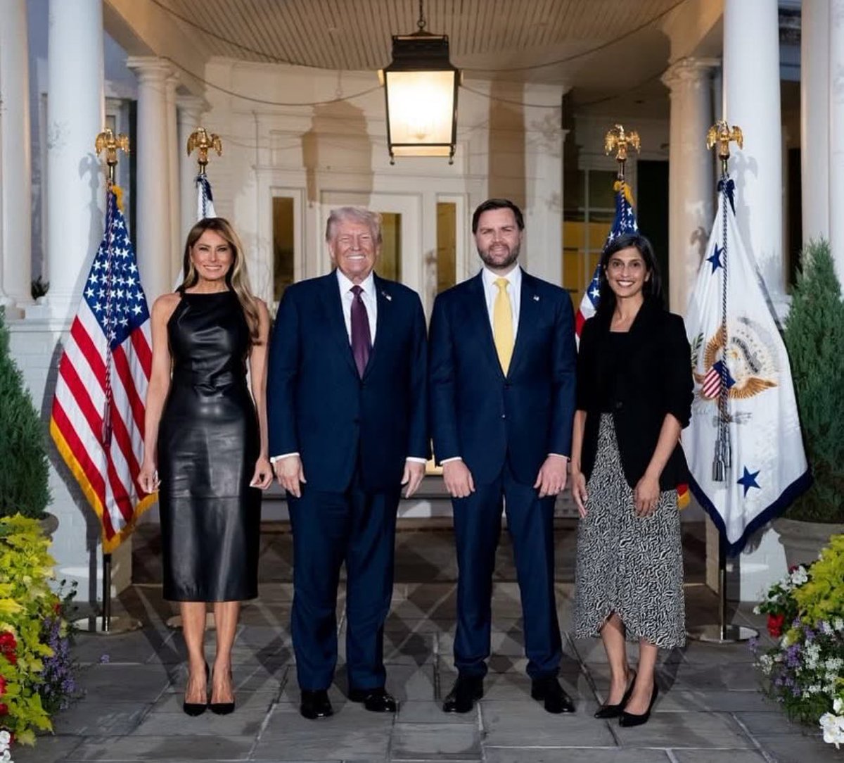 Donald Trump, Melania Trump, and two other individuals standing on a porch. American flags and presidential flags are visible on either side. The group is dressed in formal attire, including suits and dresses.