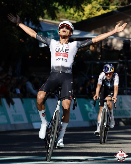 Isaac del Toro riding a bicycle, wearing a white and black UAE Team Emirates jersey with red and black accents, raising both arms in a celebratory pose. He is crossing a finish line on a road, with another cyclist in the background wearing a helmet and riding a bicycle. Text on his jersey reads "UAE Emirates" and "XFRG," and a watermark with "UAE" is visible.