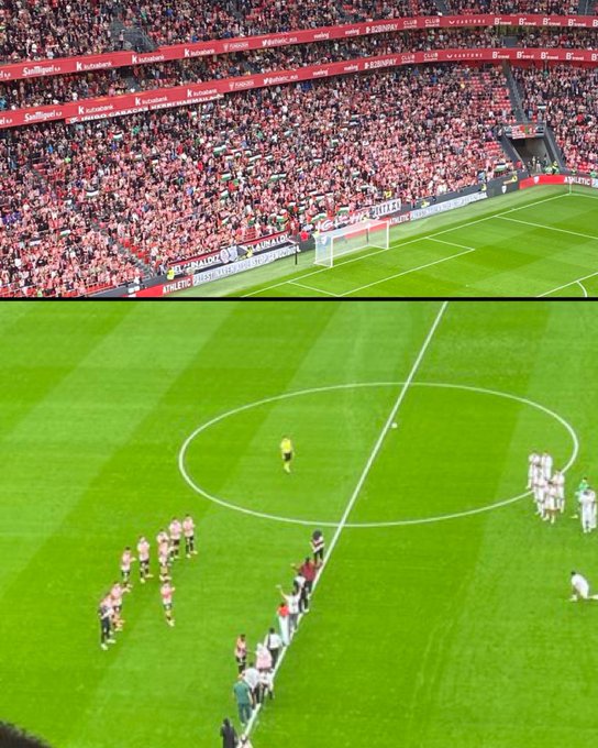 A packed stadium with thousands of spectators holding red and white flags in the stands, surrounding a soccer field. On the field, players in red and white uniforms and players in white uniforms stand near the center circle, with a referee in black. Palestinian refugees are visible among the players, and a goalpost is seen in the background.