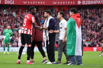 A soccer player in a red and white striped jersey with the name "Williams" and number 9 on the back shakes hands with individuals holding a Palestinian flag and wearing traditional attire on a grassy field. A crowd in the stands waves Palestinian and Athletic Bilbao flags. Banners display "Kutxabank," "Euskadi," "Athletic Club," and "STOP GENOCIDE IOA."