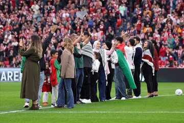 A soccer player in a red and white striped jersey with the name "Williams" and number 9 on the back shakes hands with individuals holding a Palestinian flag and wearing traditional attire on a grassy field. A crowd in the stands waves Palestinian and Athletic Bilbao flags. Banners display "Kutxabank," "Euskadi," "Athletic Club," and "STOP GENOCIDE IOA."