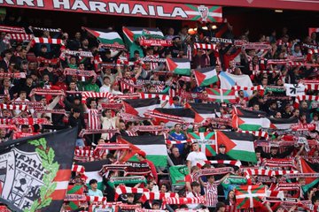 A soccer player in a red and white striped jersey with the name "Williams" and number 9 on the back shakes hands with individuals holding a Palestinian flag and wearing traditional attire on a grassy field. A crowd in the stands waves Palestinian and Athletic Bilbao flags. Banners display "Kutxabank," "Euskadi," "Athletic Club," and "STOP GENOCIDE IOA."