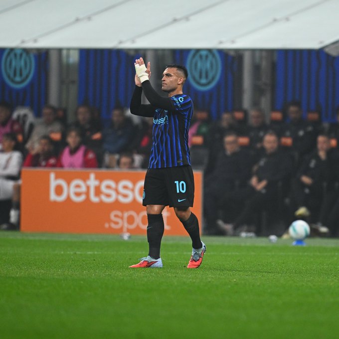 Lautaro Martinez wearing an Inter Milan jersey with the number 10, standing on a soccer field, clapping with bandaged hands. A soccer ball is visible nearby. Blue and black banners with the Inter Milan logo are in the background, along with spectators and a "betsson.sport" advertisement.