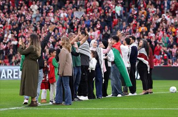 A soccer player in an Athletic Bilbao jersey with the number 9 shaking hands with individuals holding Palestinian flags on a stadium field. Fans in the stands wave Palestinian and Athletic Bilbao flags. A crowd of people, including children, stands on the field, some wrapped in Palestinian flags. Banners and text like "Digi," "Castore," and "B2BINPAY" are visible in the background.