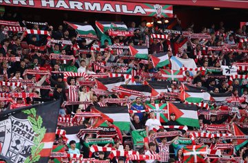 A soccer player in an Athletic Bilbao jersey with the number 9 shaking hands with individuals holding Palestinian flags on a stadium field. Fans in the stands wave Palestinian and Athletic Bilbao flags. A crowd of people, including children, stands on the field, some wrapped in Palestinian flags. Banners and text like "Digi," "Castore," and "B2BINPAY" are visible in the background.