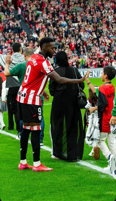 Iñaki Williams wearing an Athletic Club jersey with the number 9, shaking hands with a young boy on a soccer field. The boy wears a green and white outfit. A crowd of spectators fills the stands in the background, and a woman in a black outfit stands nearby.
