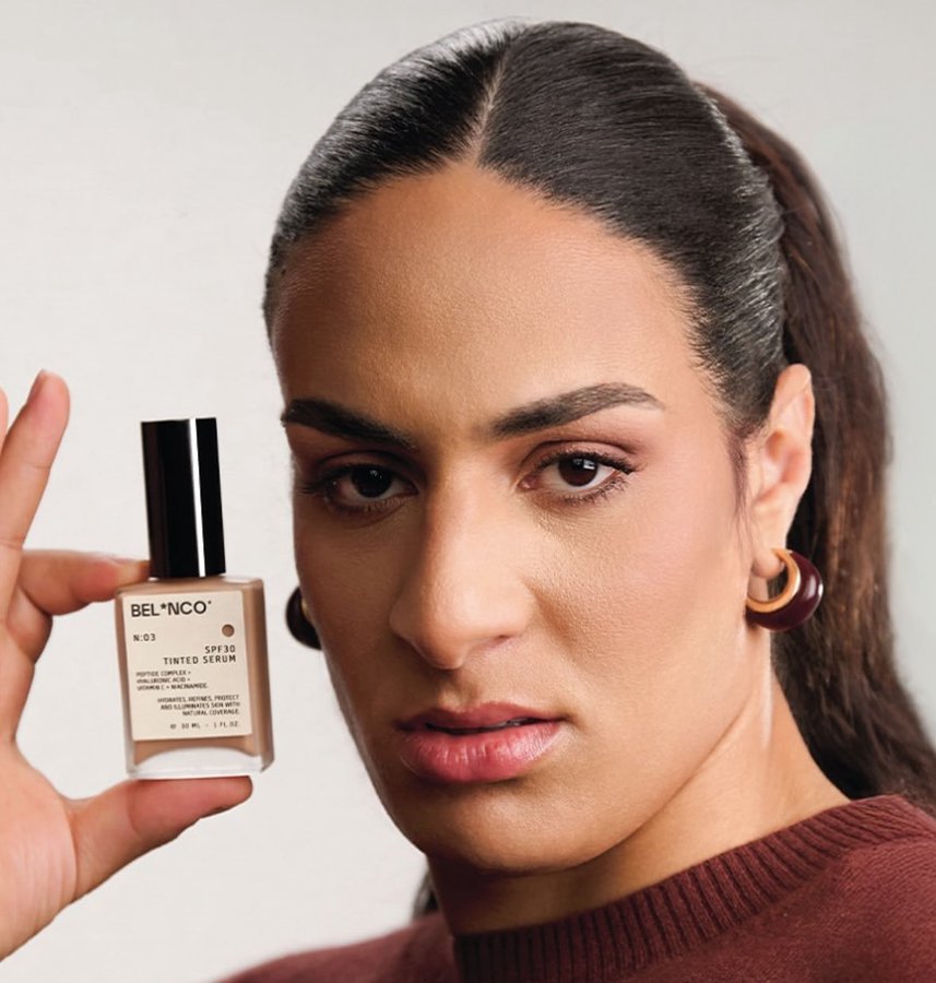 A person with long dark hair in a ponytail, wearing large hoop earrings and a brown sweater, holding a small bottle of Belnco tinted serum. The bottle has a black cap and beige label with text. The person’s face is visible, with makeup applied.