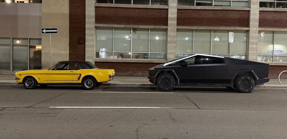 VA5SCA's tweet photo. @LSlaterPhoto @TheGuyBackstage  "Two cars in front of an office building. On the left, a canary-yellow 1966 Ford Mustang, small, sporty, looks agile and friendly. On the right, a black Cybertruck looking like Darth Vader gained 60 pounds and lost his sense of humour." https://t.co/CYJE7jYYG1
