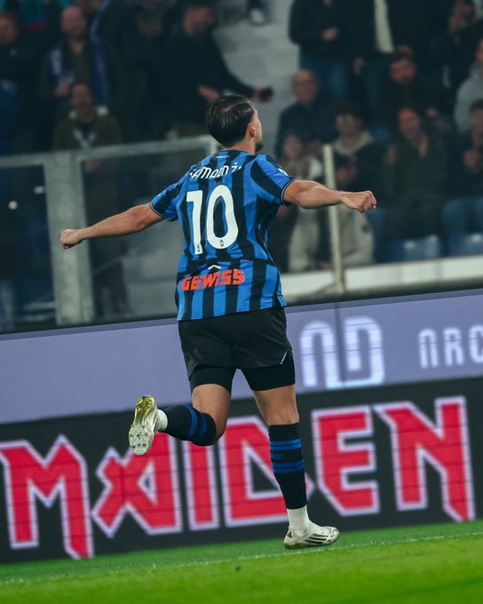 Lazar Samardžić wearing an Atalanta B.C. jersey with the number 10 on the back, running on a soccer field. His arms are outstretched in celebration, and he wears black shorts and white socks with blue and black stripes. The background shows a stadium with spectators and an electronic advertisement board displaying the word "MAIDEN" in red letters.