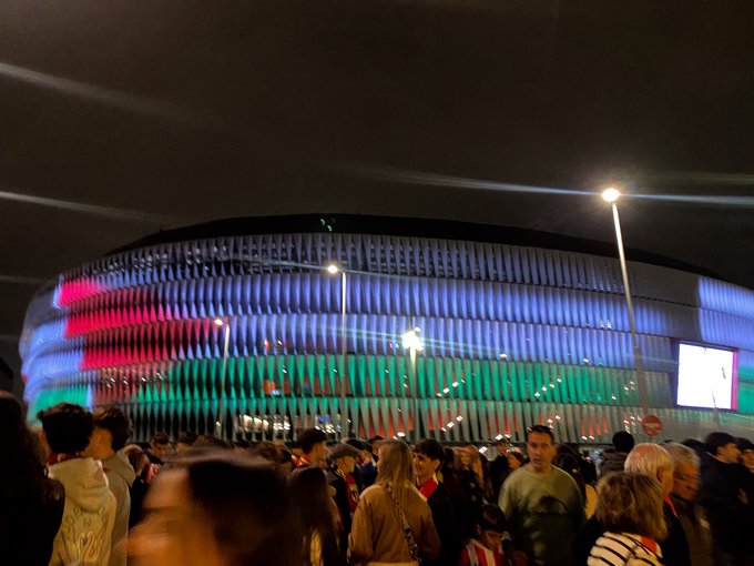 A large stadium illuminated with colorful red, green, and blue lights at night. A crowd of people stands in the foreground, some wearing casual clothing. Streetlights and a large screen are visible near the stadium.