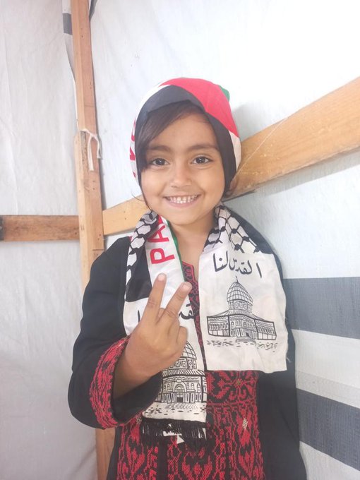 A child wearing a red and white keffiyeh with black and white checkered patterns, a black jacket, and a red patterned dress. The child makes a peace sign with their right hand. A scarf around the child\'s neck features an image of the Dome of the Rock and text in Arabic and English reading "Palestine." The background includes a wooden beam and striped fabric.