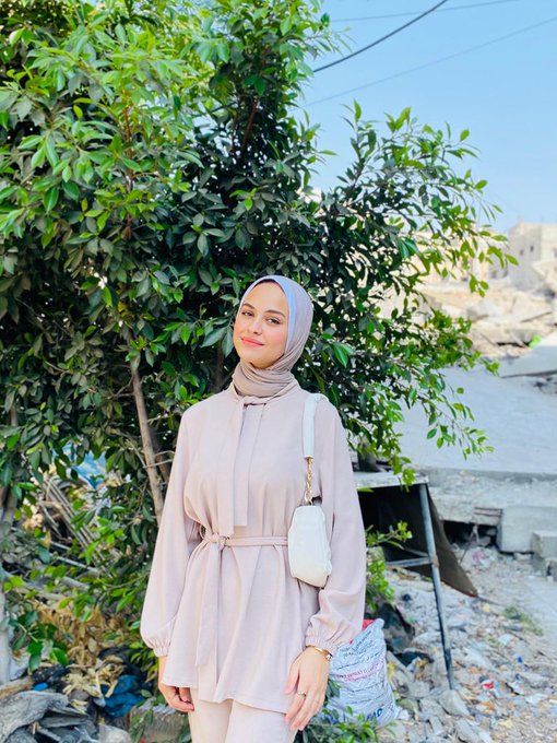Saja Al-Madhoun, a 16-year-old, stands outdoors in front of green leafy trees and a rocky background. She wears a light beige hijab and matching long-sleeved outfit with a belt, holding a white handbag over her shoulder.