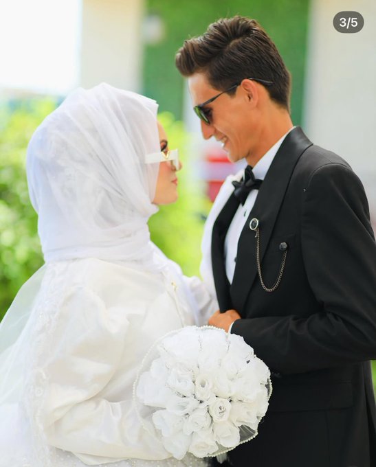 A man in a black suit and bow tie stands next to a woman in a white wedding dress and headscarf, holding a bouquet of white flowers. The setting appears to be outdoors with greenery in the background.